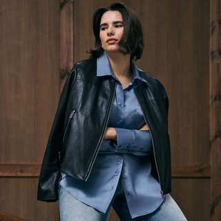 A plus-size model with short dark hair stands against a dark wood-paneled wall. She is wearing a light blue satin button-down shirt tucked into light-wash jeans, with a black faux-leather motorcycle jacket draped over her shoulders. She has her arms crossed and is looking off to the side with a calm expression.