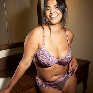 A plus-size model with long dark hair smiles broadly while leaning against a wooden vanity in a bathroom. She is wearing a purple leopard-print lingerie set, featuring a push-up bra with lace-up detail at the center and matching high-waisted briefs. She has a small floral tattoo on her inner right forearm.
