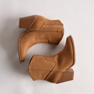 A studio product shot of a pair of tan suede ankle boots arranged on a plain light grey background. The boots feature a pointed toe, a chunky stacked block heel, and decorative western-style stitching on the sides. One boot is positioned with its side facing up, showing a matching tan zipper, while the other is placed below it facing the opposite direction.