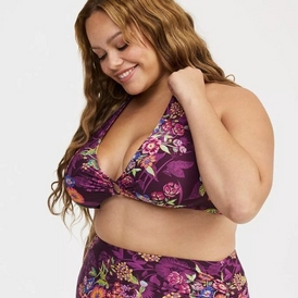 A studio portrait of a plus-size model with long, wavy blonde hair looking down and smiling against a solid white background. She is wearing a dark purple bikini featuring a vibrant floral pattern in shades of pink, yellow, and green. The bikini top has a halter-style neckline with a small ring detail at the center of the bust, and it is paired with matching high-waisted bottoms.