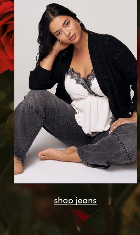A fashion photograph featuring a model with long black wavy hair and silver hoop earrings, sitting barefoot on a white surface against a light gray background. She wears a black open-knit cardigan over a white silk camisole with black lace trim at the neckline, paired with dark gray or black denim jeans. Her pose is relaxed with one hand touching her neck and head tilted slightly, looking directly at the camera with a subtle expression. The image is framed with decorative red rose petals visible on the left side and blurred floral elements in the background. At the bottom of the image, white underlined text reads 'shop jeans' against a darker background overlay.
