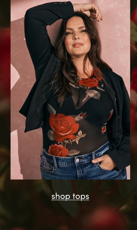 A fashion photograph featuring a model with long dark wavy hair against a soft pink textured background. She wears a black sheer mesh top with a dramatic red rose floral print pattern, layered under a black blazer with the left sleeve visible. The outfit is paired with dark blue jeans with contrast stitching. She poses with one hand raised to her hair, looking directly at the camera with a confident expression. The image is framed with blurred red rose elements on the sides. At the bottom, white underlined text reads 'shop tops' against a darker background overlay.