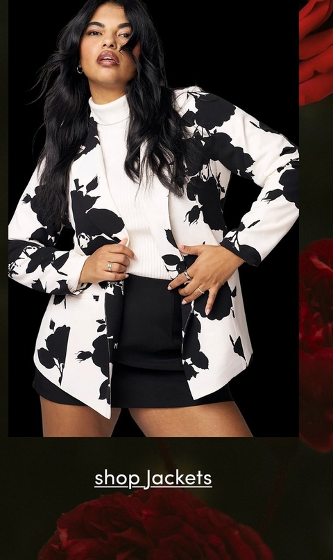 A fashion photograph featuring a model with long wavy black hair and silver hoop earrings against a solid black background. She wears a white ribbed turtleneck sweater layered under a striking white blazer with a bold black floral silhouette print, paired with black shorts. She holds a small black handbag at waist level and wears rings on her fingers. Her confident pose shows her looking upward with one hand on her hip. The image is framed with decorative red rose elements in the corners. At the bottom, white underlined text reads 'shop Jackets' against a dark background with red rose accents.