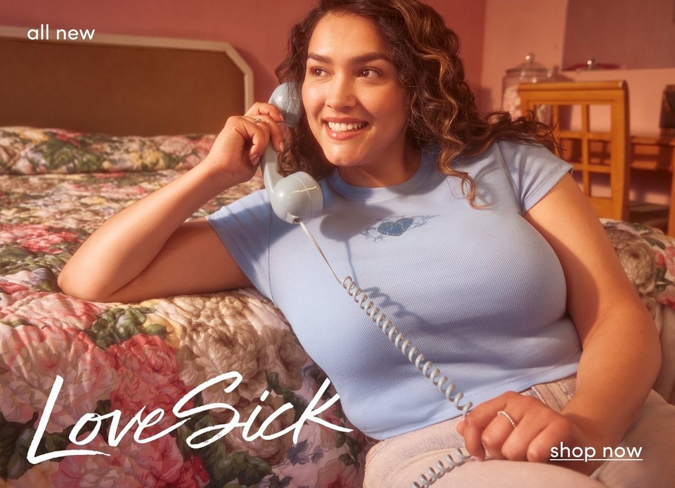 A promotional lifestyle photograph featuring a young woman with shoulder-length wavy brown hair lying on a bed with floral patterned bedding in pink, green, and cream tones. She is smiling warmly while holding a vintage-style light blue rotary telephone to her ear. She wears a fitted light blue ribbed short-sleeve top. The bedroom setting has warm coral-pink walls, a cushioned headboard visible in the background, and a wooden nightstand with glass containers. In the top left corner, white text reads 'all new'. In the bottom left, 'LoveSick' appears in white handwritten script font. In the bottom right corner, white text with an underline reads 'shop now'.