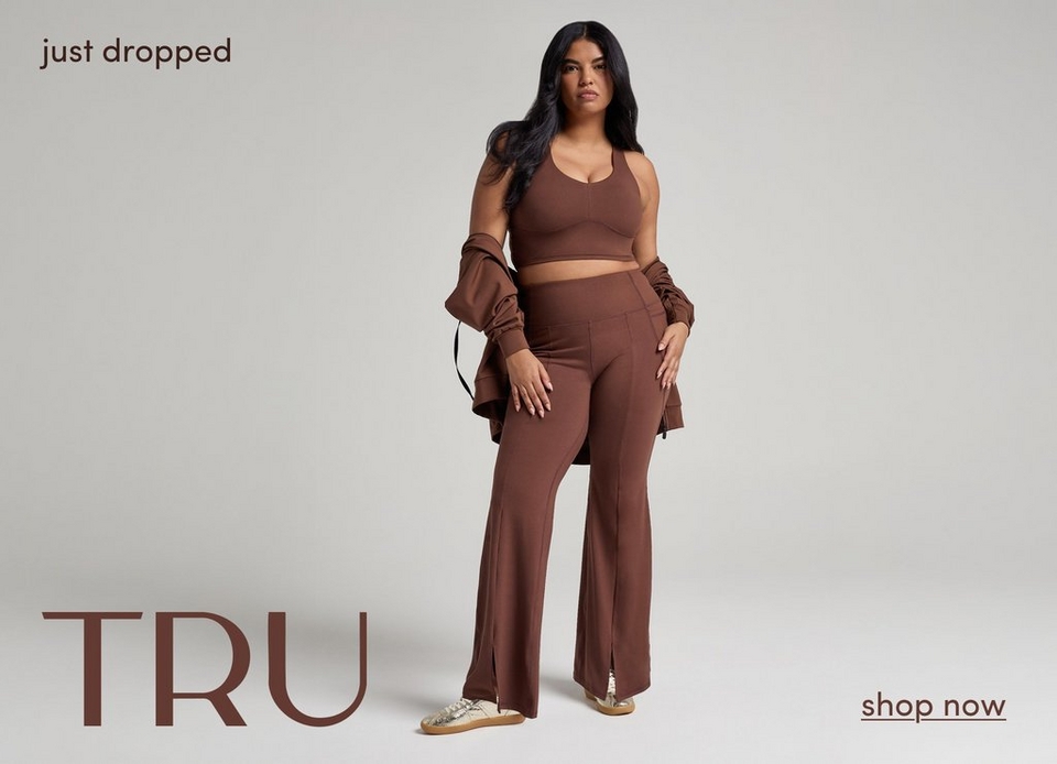 A fashion promotional photograph featuring a model with long straight black hair standing against a light gray background. She wears a coordinated chocolate brown outfit consisting of a cropped racerback tank top, high-waisted wide-leg pants, and an oversized blazer draped over her arms. The outfit is styled with beige studded slide sandals. Her pose is confident with one hand on her hip. In the top left corner, black text reads 'just dropped'. In the bottom left, large brown text displays 'TRU'. In the bottom right corner, brown text with an underline reads 'shop now'.