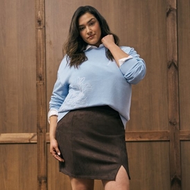A fashion photograph of a model with long dark wavy hair standing against a rustic wooden door background with visible hardware and weathered brown wood panels. She wears a light blue oversized sweatshirt with a white graphic or embroidered design on the front, paired with a black mini skirt featuring a side slit. Her pose shows her with one hand raised to her hair, looking directly at the camera with a neutral expression. The overall styling presents a casual, contemporary outfit combination.