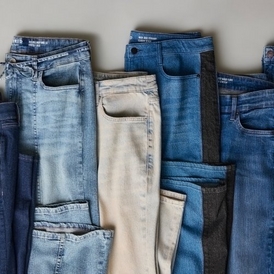 A flat lay photograph of multiple pairs of jeans arranged side by side on a light gray background. The jeans are folded to show their front sides and are displayed in various shades of denim wash, ranging from light acid wash blue on the left, to pale cream or off-white in the center, to medium and darker blue washes on the right, with what appears to be a black or very dark wash pair also visible. The jeans show typical denim details including front pockets, waistbands, and button flies. The arrangement creates a gradient effect from light to dark denim tones.