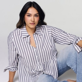 A fashion photograph of a model with long dark hair with lighter highlights, wearing silver hoop earrings. She is dressed in an oversized purple and white vertical striped button-up shirt with the front partially unbuttoned and tied at the waist, paired with light blue jeans. She sits in a relaxed pose against a light gray background, with one arm resting on her bent knee. Her expression is neutral and she looks directly at the camera. The styling presents a casual, effortless aesthetic.