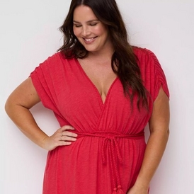 A fashion photograph of a smiling model with long dark brown hair looking down, photographed against a light gray background. She wears a bright coral-red dress with a wrap-style v-neckline, short pleated cap sleeves, and a drawstring waist tie. The dress appears to be made of a soft, textured knit fabric. She poses with one hand on her hip in a relaxed, joyful stance. The image is cropped to show from approximately mid-thigh up.