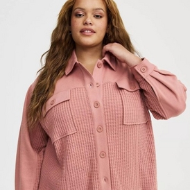 A fashion photograph of a model with long wavy brown hair against a light gray background. She wears an oversized dusty rose pink shirt jacket featuring a textured waffle-knit pattern on the front panels, smooth fabric on the collar and button placket, two chest pockets with flaps, and long sleeves with button cuffs. The garment has a relaxed, oversized fit and buttons down the front. She poses with one hand raised to her hair, looking directly at the camera with a neutral expression. The image is cropped to show from approximately mid-torso up.