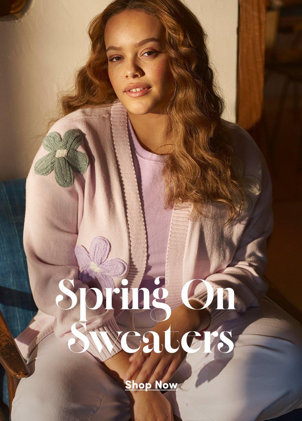 A woman sits in warm sunlight against a cream-colored wall with wooden trim visible in the background. She wears a dusty pink knit hoodie with ribbed drawstring hood and cuffs, featuring embroidered floral appliques in sage green and lavender on the sleeves and front. The hoodie has a kangaroo pocket. She pairs it with light-colored pants and sits on what appears to be a blue cushioned surface. She has long wavy honey blonde hair and is looking directly at the camera. In the center of the image is white serif text reading 'Spring On Sweaters' and below it 'Shop Now' with an underline.