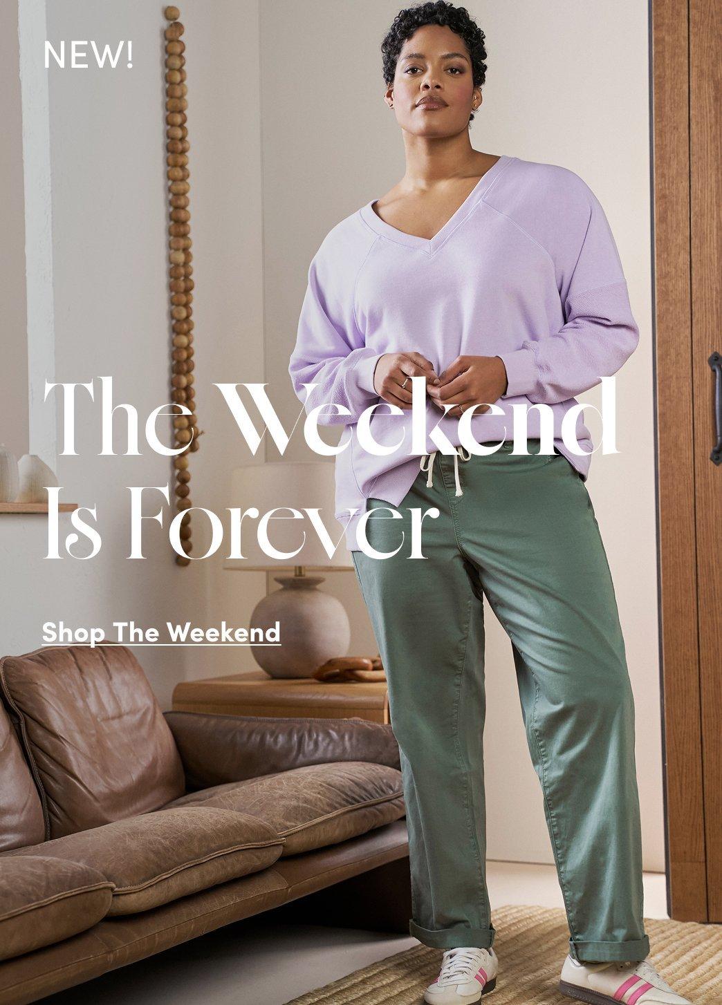 A woman stands in a minimalist living room with white walls, wooden trim, and a tan leather sofa. She wears a lavender long-sleeve V-neck sweatshirt and sage green wide-leg pants with a white drawstring waist, paired with white sneakers with pink stripes. She has short dark curly hair and small hoop earrings. Her hands are clasped at her waist. Behind her is a wooden shelf with a decorative ceramic vase and a wooden bead garland hanging on the wall. She stands on a natural woven jute rug. In the upper left corner is white text reading 'NEW!' In the center is large white serif text reading 'The Weekend Is Forever' and below it 'Shop The Weekend'.