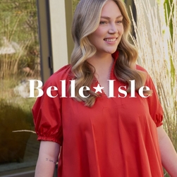 A woman stands outdoors smiling against a background of natural foliage, greenery, and textured wall panels. She wears a bright red dress with short puff sleeves and a collared V-neckline. She has long wavy blonde hair with highlights and a small tattoo visible on her left arm. Across the center of the image is the 'Belle*Isle' logo in large white serif font with a star between the words.