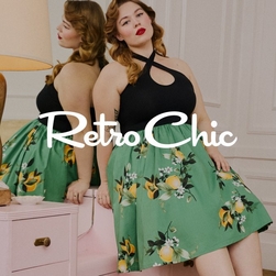 A woman poses in a vintage-styled room with cream walls and pink wainscoting, sitting on a pink dresser with gold knobs next to a large round mirror and gold table lamp. She wears a black halter-neck top with a crisscross design at the neckline paired with a sage green A-line skirt featuring a vintage lemon and floral print with yellow fruit, white blossoms, and green leaves. She has wavy honey blonde hair styled in vintage waves, red lipstick, and visible tattoos on her arms. Her reflection is visible in the mirror behind her. Across the center of the image is 'Retro Chic' in large white script font.