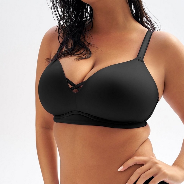 'Close-up product photo showing woman from shoulders to waist wearing black wireless bra with crisscross strap detail at center front against white background. Long dark hair visible falling over left shoulder and chest. Left hand with light nail polish rests on hip. Photo cropped at neck level.'