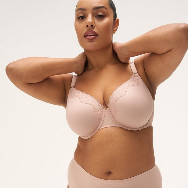 'Product photo of woman with hair slicked back wearing light pink underwire bra with scalloped lace trim along top edge of cups against light beige background. She faces forward with both hands raised behind her head, elbows out to sides. Small stud earring visible in left ear. Matching light pink underwear visible at bottom of frame.'