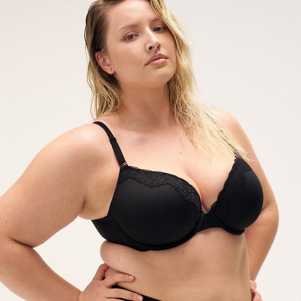 'Product photo of woman with long blonde hair wearing black underwire bra with lace trim detail along top edge of cups against light beige background. She faces forward with head tilted slightly upward and to her right. Small stud earrings visible in both ears. Right hand rests on hip, left arm out of frame.'