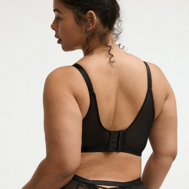 'Back view product photo of woman with dark brown hair pulled up in messy bun wearing black bra against light beige background. She faces away showing back closure with hook-and-eye fastening, U-shaped back panel, and adjustable straps. Matching black underwear visible at bottom of frame.'