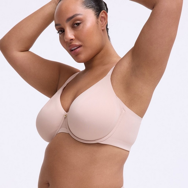 'A plus-size model poses against a light gray background with both arms raised above her head. She wears a smooth full-coverage underwire bra in a light nude blush color with a small gold front hardware detail. She has her dark hair pulled back and looks toward the camera.'