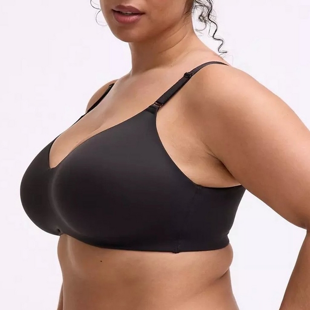 'Close-up product photo showing woman from chin to waist wearing black wireless bra with V-neckline and adjustable straps against light pink background. Dark hair pulled up with loose strands visible. Photo cropped at chin level, showing front three-quarter view angled slightly to left.'