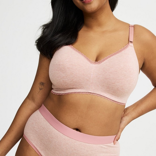 'Product photo of woman with long dark hair wearing light pink wireless bra with lace trim along top and bottom edges and adjustable straps against light beige background. She faces forward with right hand on hip. Photo cropped at chin level. Small floral tattoo visible on left upper arm. Matching light pink heathered underwear with pink waistband visible at bottom of frame.'
