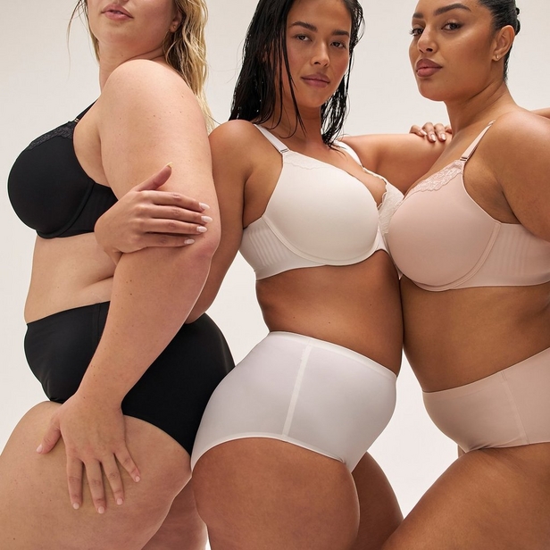 'Group photo of three women standing together against light beige background, each wearing different colored bra and matching underwear. Left: woman with long blonde hair wearing black underwire bra with lace trim and black high-waist underwear, body angled to her left showing right side, right hand rests on hip. Center: woman with long dark hair wearing ivory or off-white underwire bra with textured cups and white high-waist underwear, facing forward. Right: woman with dark hair pulled back wearing light beige underwire bra with lace trim and matching beige high-waist underwear, body angled to her right, left hand rests on center woman shoulder. Gold necklace visible on right woman. All three cropped at chin level.'