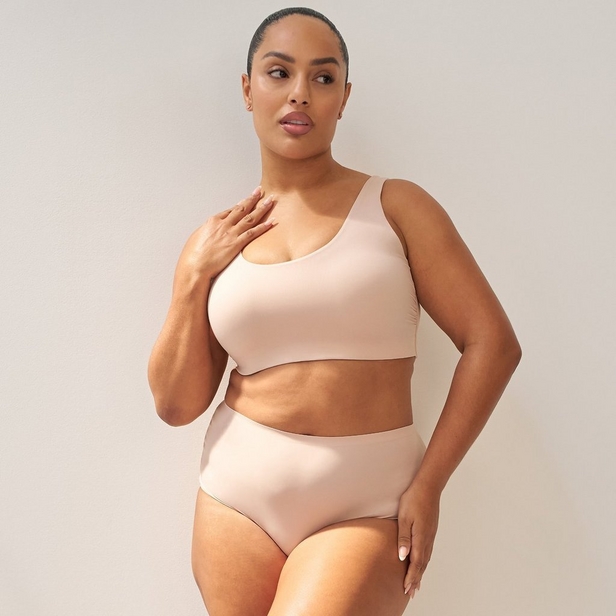 'Product photo of woman with dark hair slicked back wearing light beige wireless bra with scoop neckline and wide shoulder straps against light beige background. She faces forward with left hand raised to chest near collarbone, right hand on hip. Small stud earrings visible in both ears. Matching light beige high-waist underwear visible at bottom of frame.'