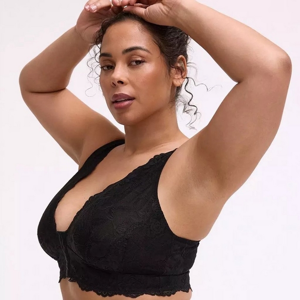 'Product photo of woman with curly dark hair pulled back wearing black lace wireless bralette with V-neckline and adjustable straps against light pink background. She faces forward with both arms raised above her head, hands adjusting her hair. Small stud earring visible in left ear.'