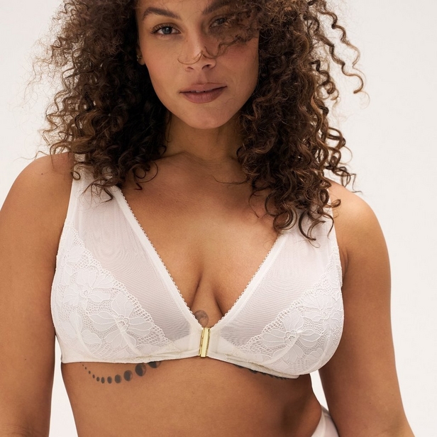 'Product photo of woman with curly brown hair wearing ivory or off-white lace underwire bra with front clasp closure against light beige background. She faces forward, hair falling over both shoulders. Small tattoo visible on sternum below bra closure. Curved line tattoo visible on left ribs below bra band.'