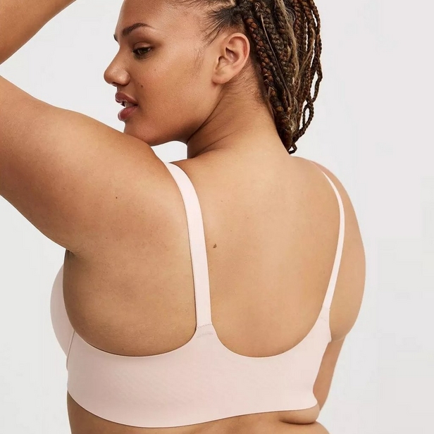 'Back view product photo of woman with braided hair pulled back wearing light pink wireless bra against light gray background. She faces away showing back panel with thin vertical center seam and adjustable straps. Left arm raised above head, right hand rests on hip out of frame. Photo cropped at back of head showing ear and profile.'
