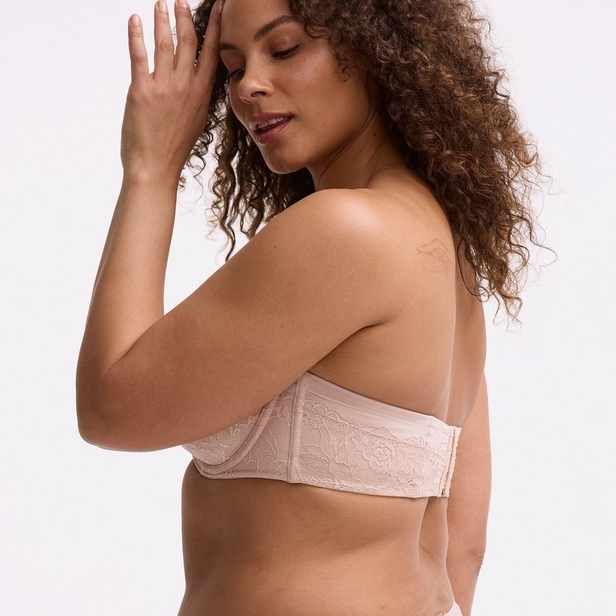 'Side view product photo of woman with curly brown hair wearing light pink or nude strapless bra with lace panel on side against light pink background. She faces to her right with left hand raised to her head near temple, right arm crosses body out of frame below chest. Hair flows over left shoulder and down back. Photo cropped at nose level.'