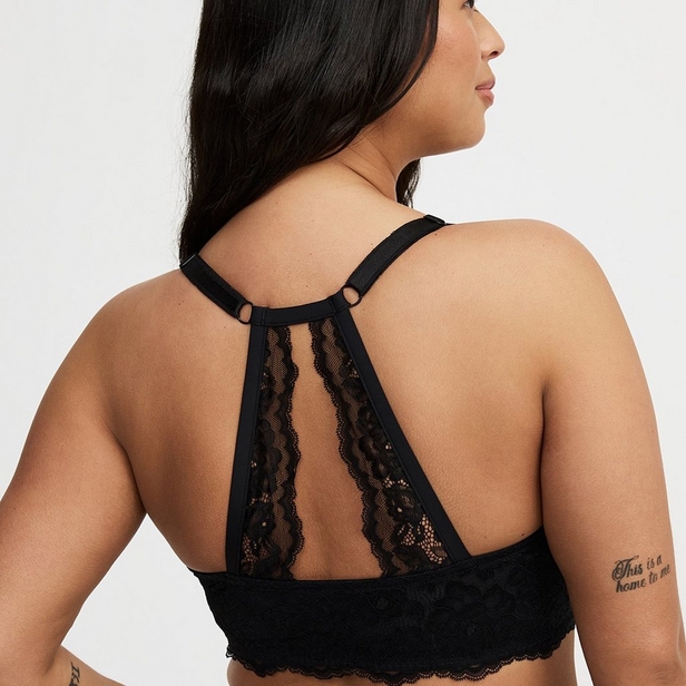 'Back view product photo of woman with long dark hair wearing black lace bralette with racerback design against light beige background. She faces away showing back panel with sheer lace inserts creating vertical stripes and scalloped lace trim along bottom edge. Two small metal rings connect racerback straps at upper back. Text tattoo visible on right upper arm reading 'This is not home to me' and decorative tattoo visible on left forearm. Photo cropped at back of head showing profile.'