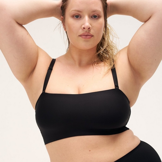 'Product photo of woman with blonde hair pulled back wearing black wireless bra with straight neckline and adjustable straps against off-white background. She faces forward with both arms raised above head, elbows bent outward. Small stud earring visible in left ear. Matching black underwear visible at bottom of frame.'