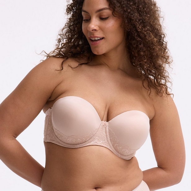 'Product photo of woman with curly brown hair wearing light pink or nude strapless bra with lace side panels against white background. She faces forward with head tilted down looking to her right, slight smile. Both arms at sides, left hand on hip. Curly hair flows over both shoulders. Photo cropped at forehead level.'