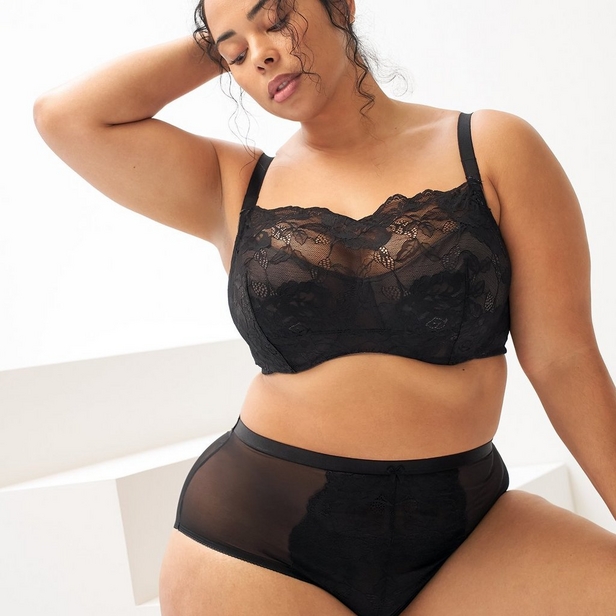 'Product photo of woman with curly dark hair wearing black lace wireless bralette with full lace coverage, scalloped trim along top edge, and adjustable straps against white background with white geometric surface visible at bottom. She sits or reclines with head tilted down and to her left, left hand raised to back of head near ear, right arm out of frame. Photo cropped at forehead level. Black sheer mesh high-waist underwear with lace trim detail visible at bottom of frame.'