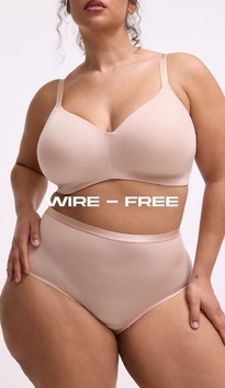Model shown from neck to mid-thigh against a light gray background, wearing a nude pink wire-free bra with wide straps and smooth cups, paired with matching high-waisted nude pink briefs with a satin waistband. Model has curly brown hair visible at shoulders and one hand resting on hip. White text overlay reads 'WIRE - FREE' across the midsection.