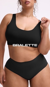 Model shown from chin to mid-thigh against a light gray background, wearing a black scoop-neck bralette with wide straps and smooth fabric, paired with matching black high-waisted briefs. Model has curly brown hair visible at shoulders, one arm raised with hand near shoulder and white nail polish visible. White text overlay reads 'BRALETTE' across the midsection.'
