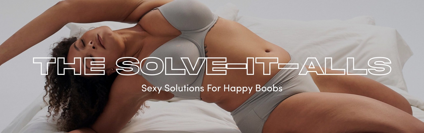 'Banner featuring woman with curly dark hair lying on white bedding wearing gray underwear set. Her left arm is raised above her head and right arm rests across her body. Large white outlined text reads 'THE SOLVE-IT-ALLS' across the center. Below in smaller white text is 'Sexy Solutions For Happy Boobs'. Light gray background visible in upper right corner.'