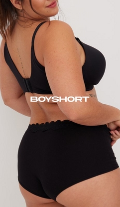 Model shown from chin to upper thighs in a back-angled view against a light gray background, wearing a black bra with smooth cups and adjustable straps visible from behind, paired with black boyshort panties featuring a scalloped lace waistband detail. Model has long straight reddish-brown hair and tattoos visible on her upper arm and forearm, with hands clasped in front. White text overlay reads 'BOYSHORT' across the midsection.