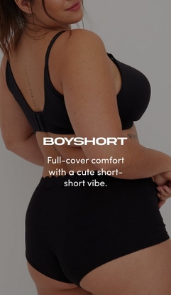 Model shown from chin to upper thighs in a back-angled view against a light gray background, wearing a black bra with smooth cups and adjustable straps visible from behind, paired with black boyshort panties featuring a scalloped lace waistband detail. Model has long straight reddish-brown hair and tattoos visible on her upper arm and forearm, with hands clasped in front. White text overlay reads 'BOYSHORT' with subtext below reading 'Full-cover comfort with a cute short-short vibe.'