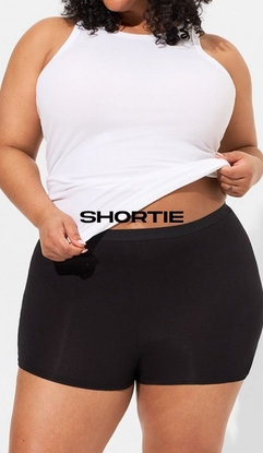 Model shown from shoulders to mid-thighs against a light gray background, wearing a white ribbed high-neck tank top lifted slightly to show the waistband, paired with black shortie underwear with a smooth waistband and short-length legs. Model has curly dark hair and is holding the hem of the tank top with both hands. Black text overlay reads 'SHORTIE' across the midsection.