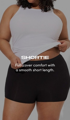 Model shown from shoulders to mid-thighs against a gray background, wearing a white ribbed high-neck tank top lifted slightly to show the waistband, paired with black shortie underwear with a smooth waistband and short-length legs. Model has curly dark hair and is holding the hem of the tank top with both hands. White text overlay reads 'SHORTIE' with subtext below reading 'Full-cover comfort with a smooth short length.'