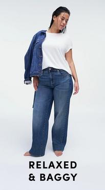 A woman with dark hair in a wet-look style and striped earrings stands barefoot against a white background, looking down. She wears a white short-sleeve crew neck tee with a medium wash wide-leg denim jacket draped over one shoulder. Bold black text at the bottom reads 'Relaxed & Baggy'.