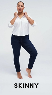 A woman with her dark hair pulled back stands barefoot against a white background, both hands raised to the collar of her shirt. She wears a white V-neck button-front blouse with rolled sleeves and dark wash skinny jeans. Bold black text at the bottom reads 'Skinny'.