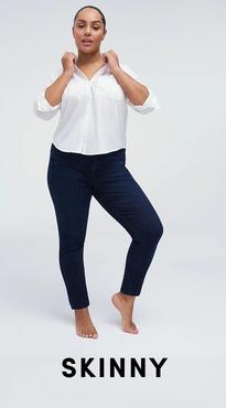 A woman with her dark hair pulled back stands barefoot against a white background, both hands raised to the collar of her shirt. She wears a white V-neck button-front blouse with rolled sleeves and dark wash skinny jeans. Bold black text at the bottom reads 'Skinny'.