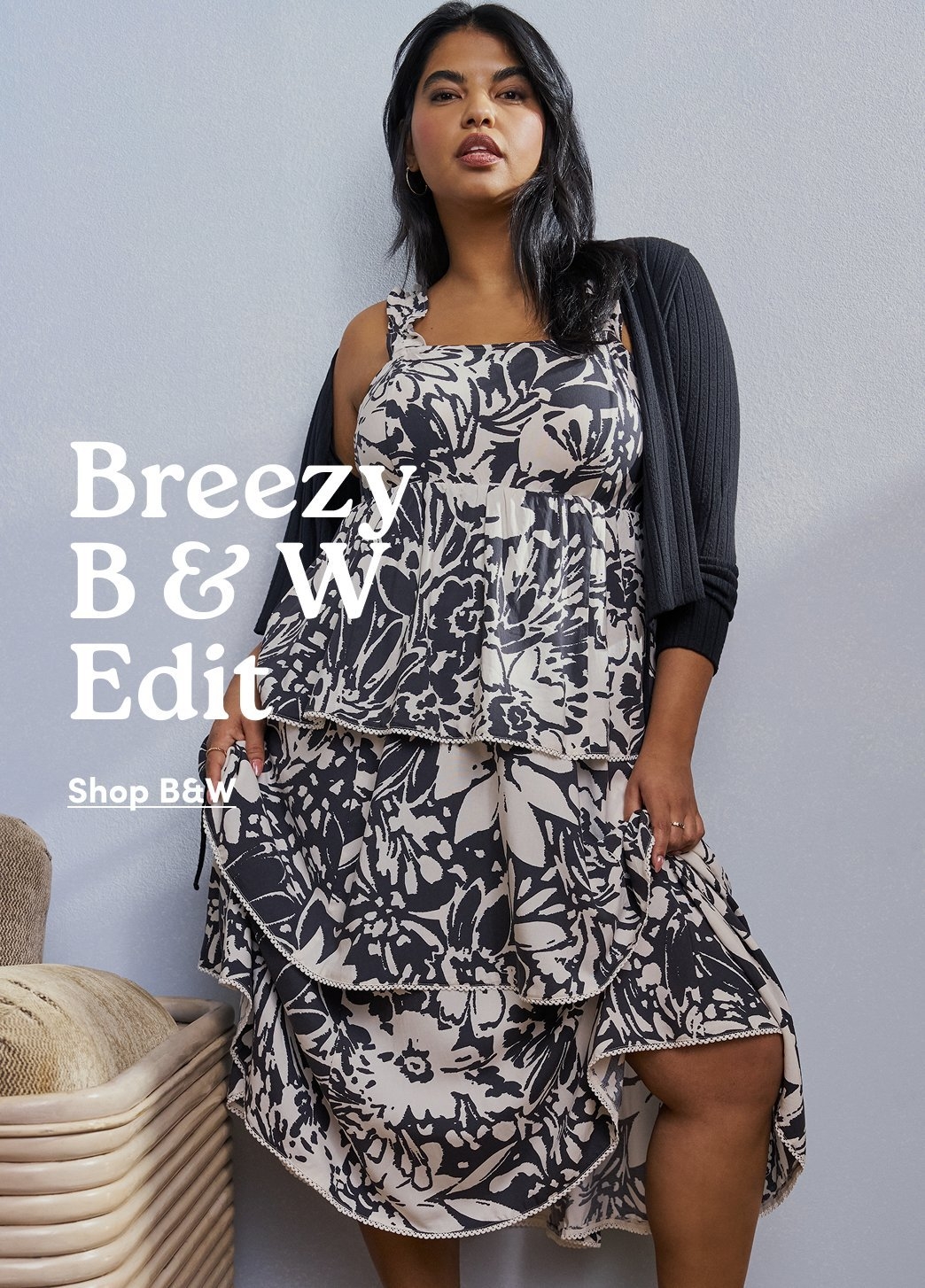 Promotional image of a model wearing a black and cream floral tiered dress with a black cardigan against a light gray wall. Text reads “Breezy B & W Edit” with “Shop B&W” below.