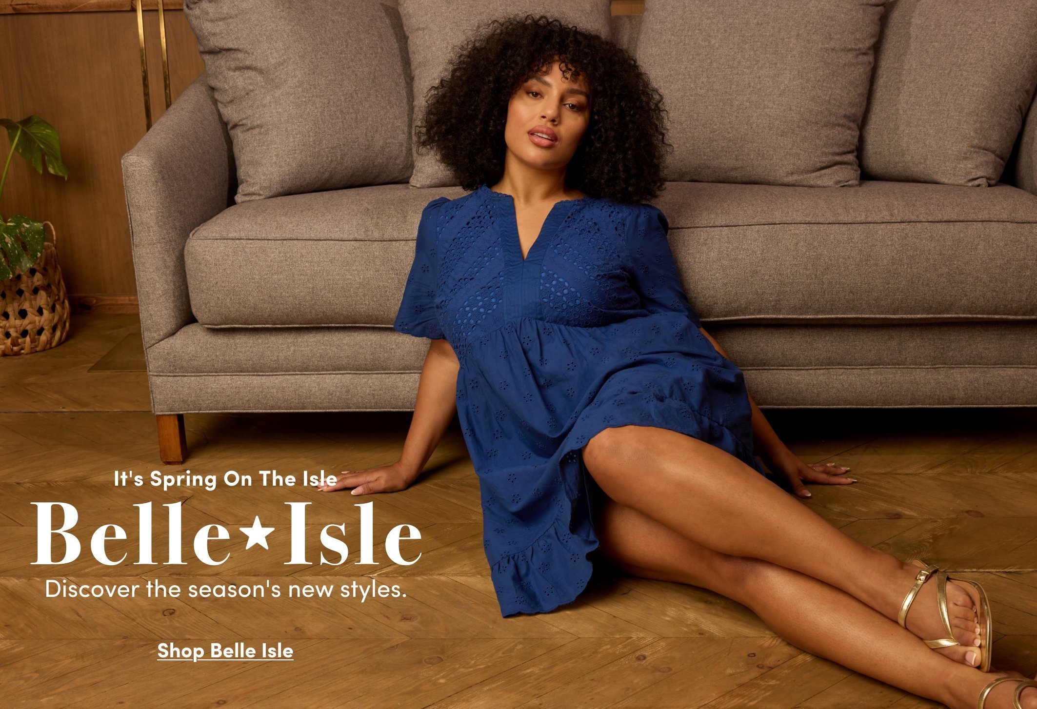 Promotional image for Belle Isle. A model reclines on a wooden floor in front of a gray sofa wearing a navy eyelet short-sleeve dress and gold sandals. Text reads “It’s Spring On The Isle Belle Isle. Discover the season’s new styles.” with “Shop Belle Isle” below.