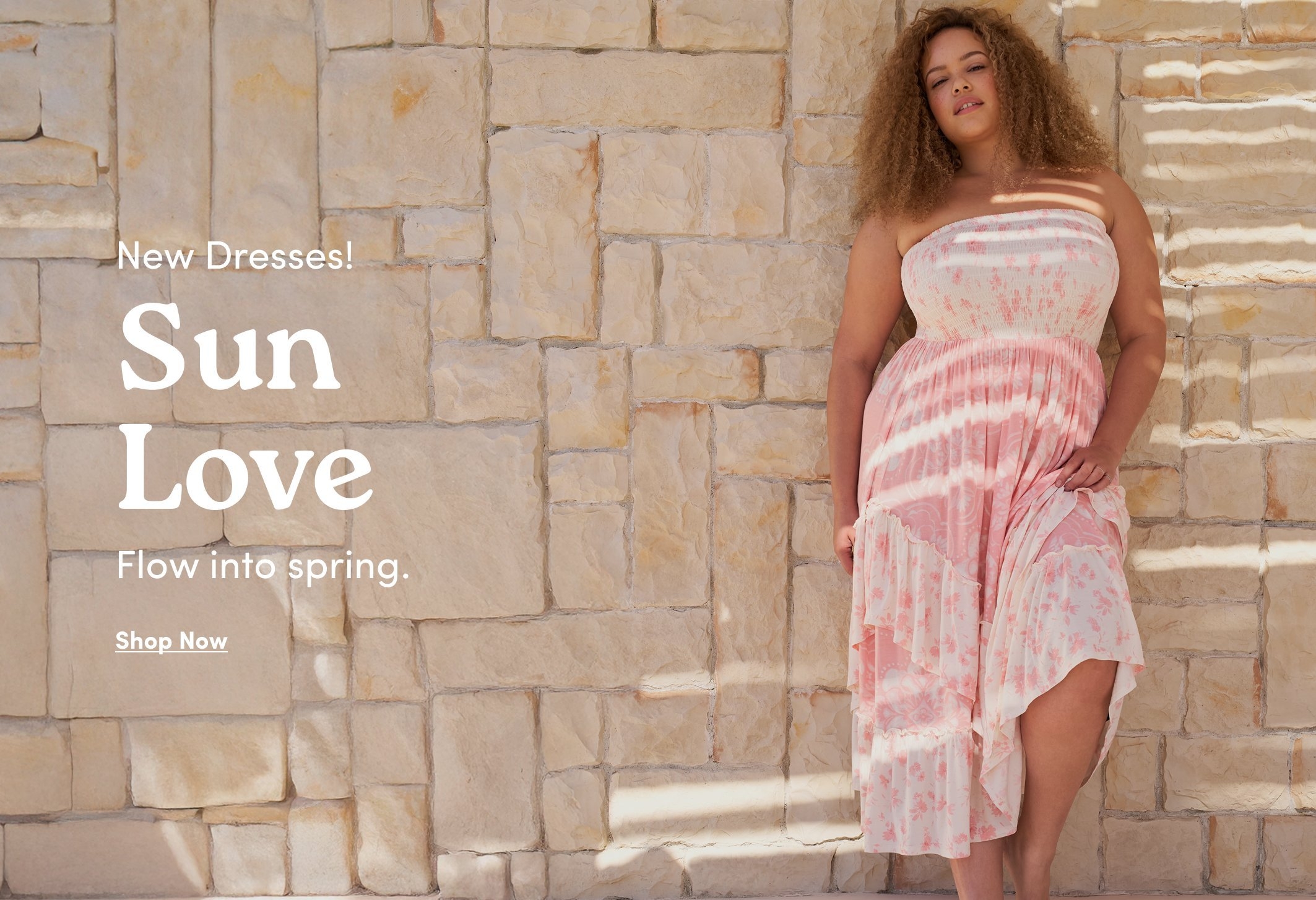 Promotional image featuring a model wearing a pink strapless floral maxi dress with a tiered skirt, standing against a light stone wall. Text reads “New Dresses! Sun Love. Flow into spring.” with “Shop Now” below.