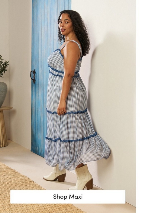 'A tall promotional banner featuring a plus-size model standing sideways and looking over her shoulder against a white wall with a weathered blue wooden door and a small green plant and gray vase visible to the left. She wears a blue and white gingham check sleeveless tiered maxi dress with blue ruffle trim detailing at the neckline and between tiers, and white block-heel western ankle boots. She has long curly dark hair. At the bottom is a white rectangular button with dark text reading Shop Maxi.'