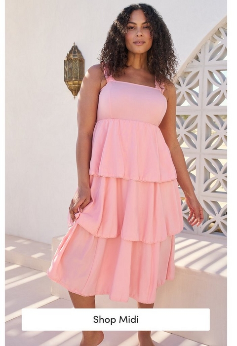'A tall promotional banner featuring a plus-size model standing outdoors on white steps against a white wall with a decorative geometric lattice screen and a gold ornate lantern visible behind her. She wears a light pink sleeveless tiered midi dress with ruffle straps and scalloped lace trim between tiers. She has voluminous curly dark hair. At the bottom is a white rectangular button with dark text reading Shop Midi.'